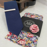 Navy tie with silver tie bar, floral pocket square, and a pink and white flower lapel pin.